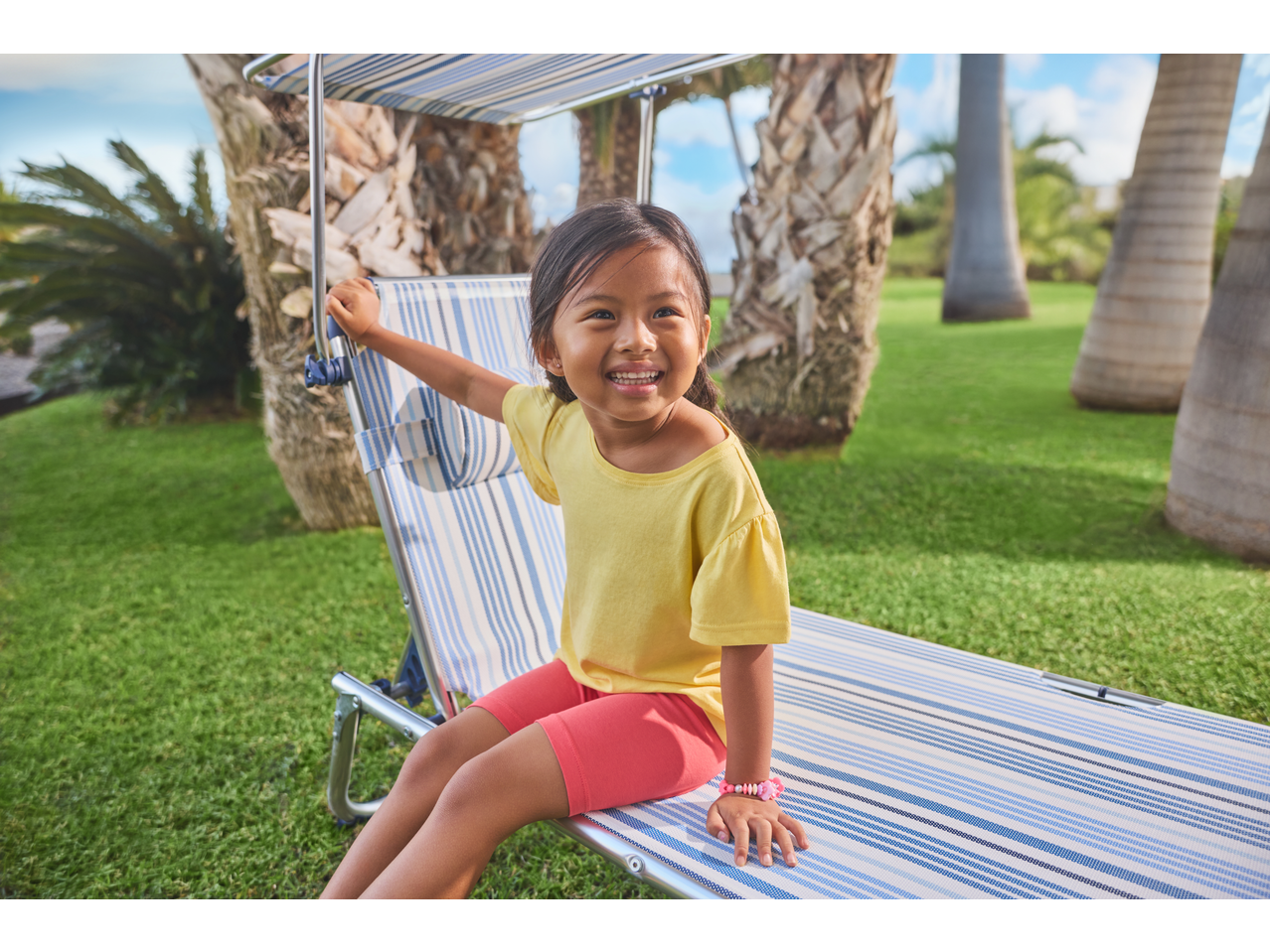 Menina com leggings Capri LUPILU amarelas e cor-de-rosa, sorrindo numa espreguiçadeira de jardim.