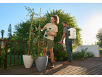 Woman tending to potted tomato plants, man spraying flowers with a backpack sprayer.