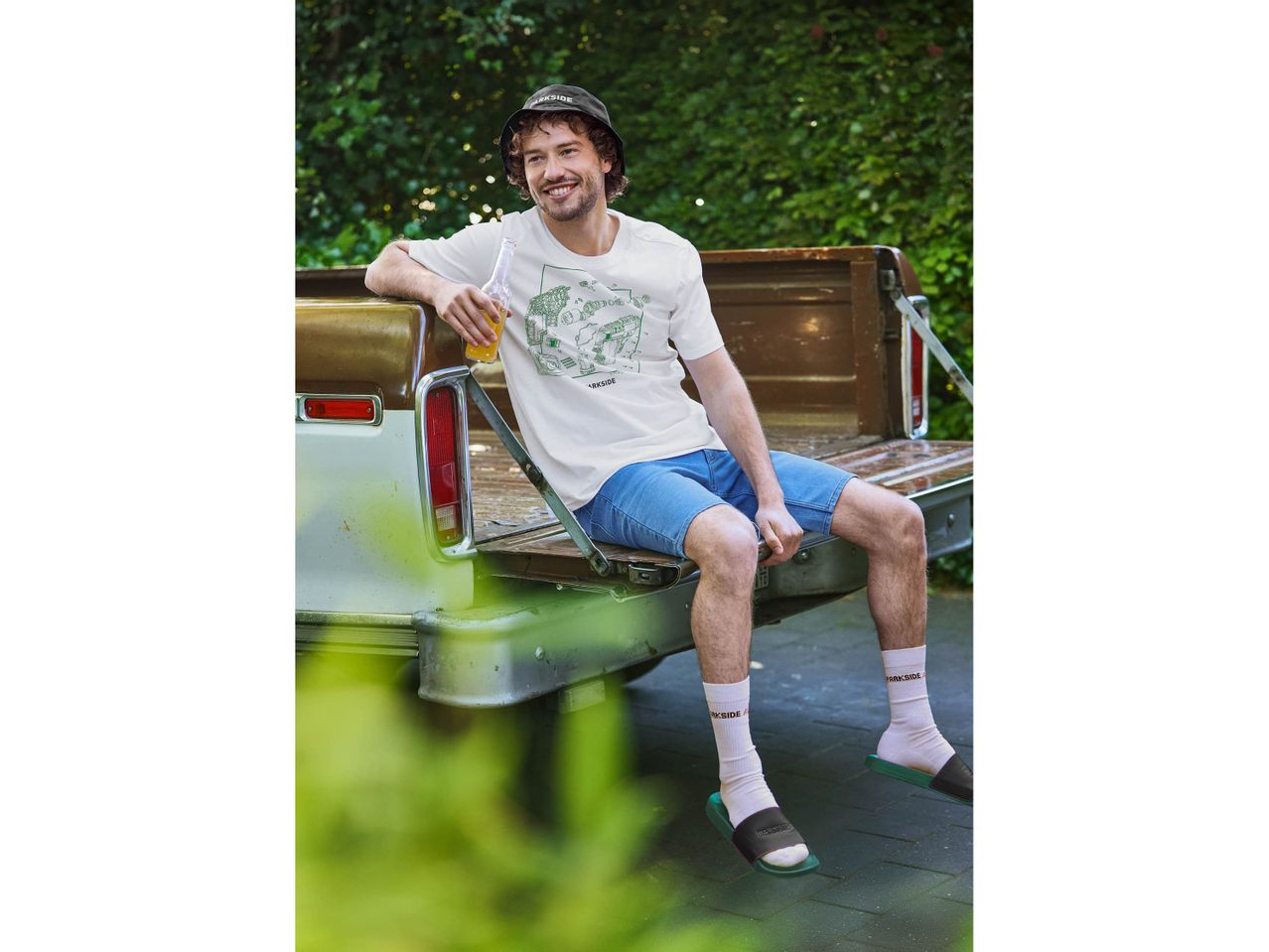 Man in Parkside bucket hat, t-shirt, shorts, socks, and slides, sitting on a truck tailgate.