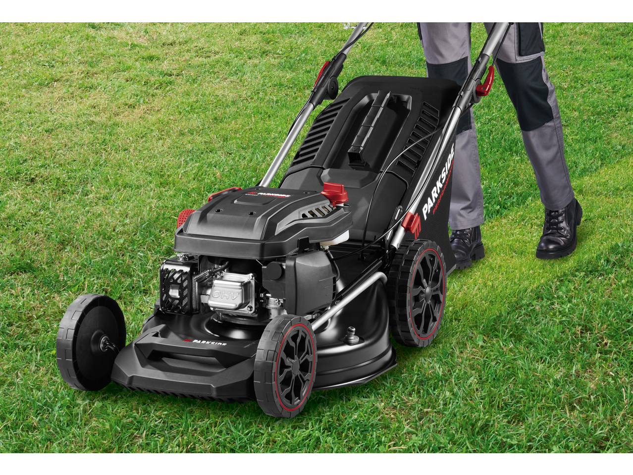 Person in work trousers pushing a black lawnmower with red accents on green grass.
