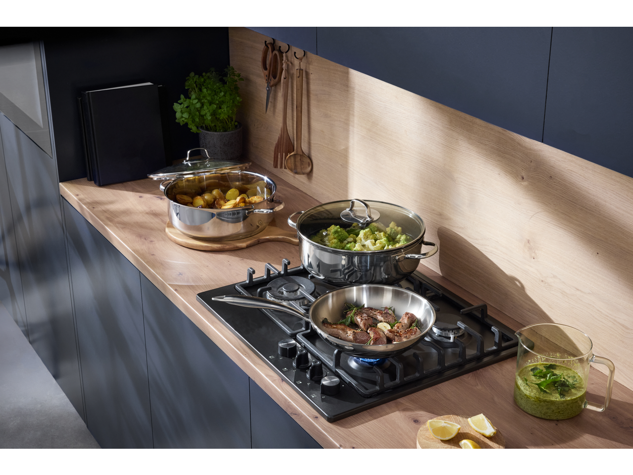 Bucătărie modernă cu plită pe gaz, tigaie cu carne, oală cu broccoli și cartofi prăjiți.