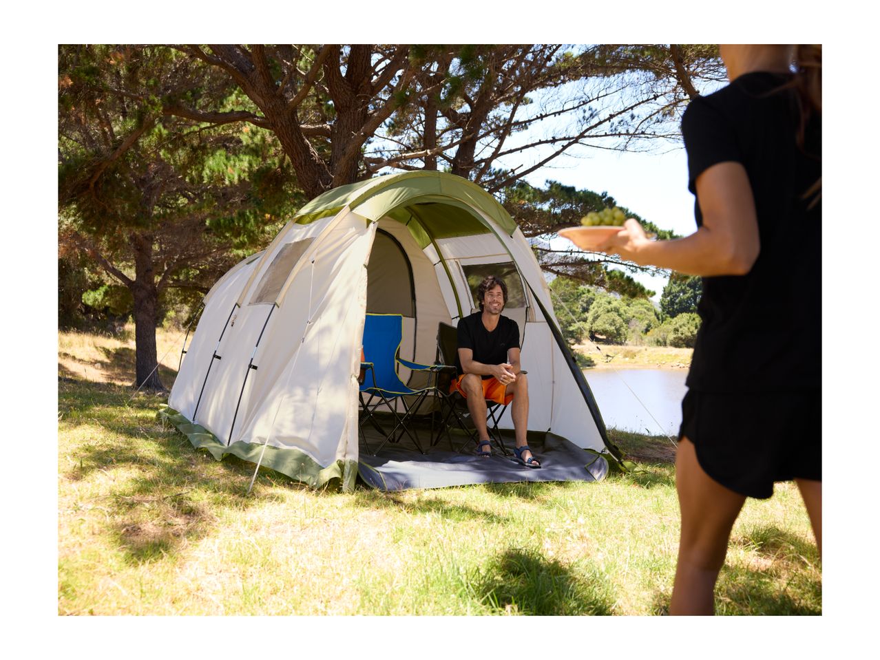 Man sitting in a camping tent, being offered grapes by a woman, by a lake.