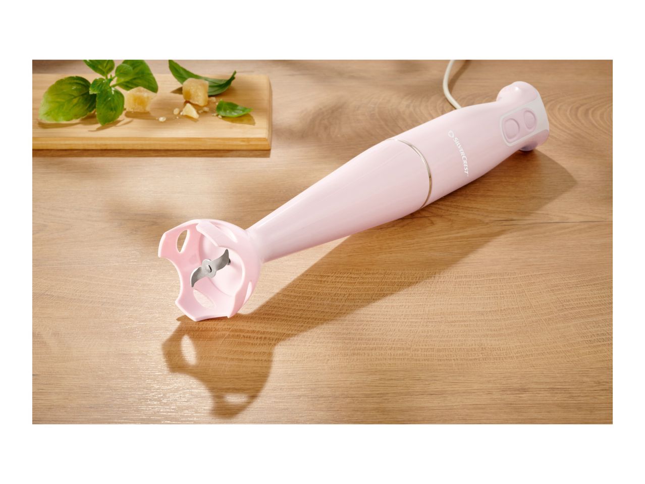 A pink SilverCrest immersion blender on a wooden surface next to basil and cheese.
