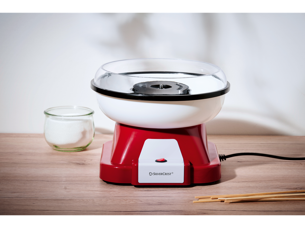 Crvena Silvercrest mašina za šećernu vunu sa posudom šećera i štapićima na drvenom stolu.