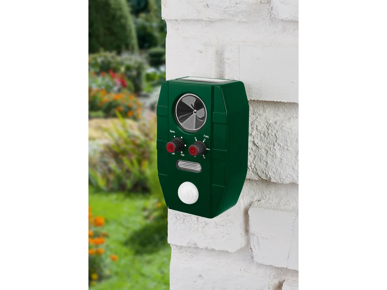 Green ultrasonic animal repeller with solar panel mounted on a white brick wall in a garden
