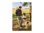 Man in a green bomber jacket and cargo pants interacting with a dog by a fence in a field.