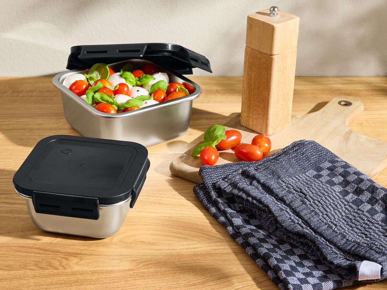 Ανοξείδωτα δοχεία φαγητού με σαλάτα καπρέζε, ξύλινος μύλος πιπεριού και πετσέτα.