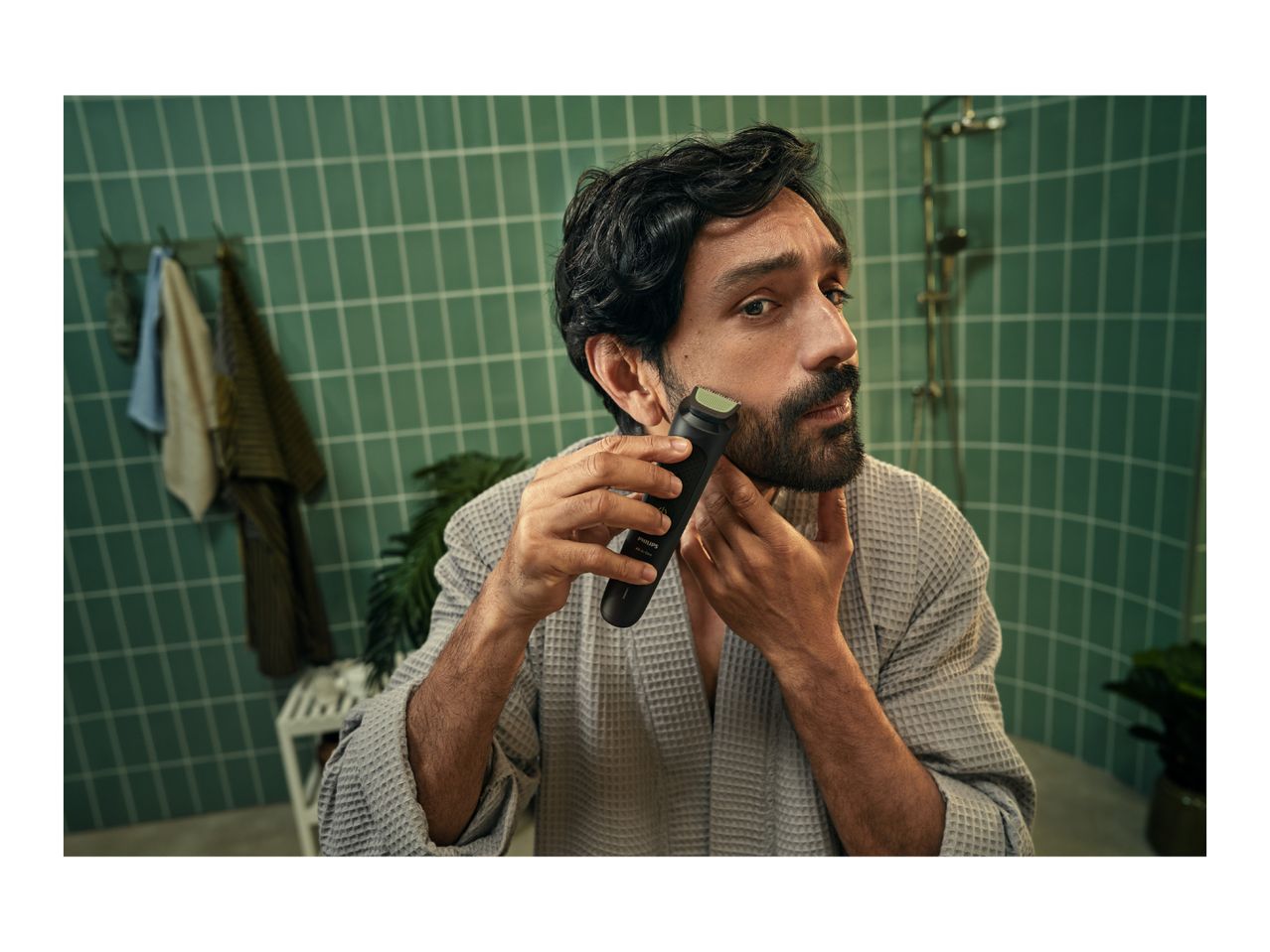 Man in a bathrobe trimming his beard with an electric shaver in a bathroom with green tiles.