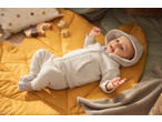 Happy baby in a grey hooded romper lying on a yellow quilted play mat with toys.