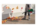 Person using a leaf blower to clear autumn leaves from a paved area next to a white wall.