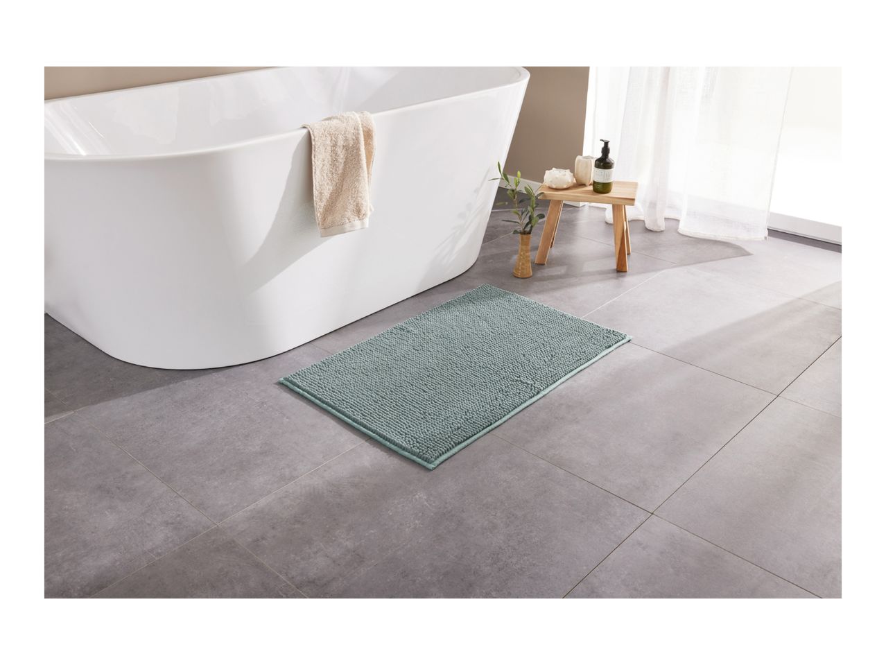 A light green bath mat on a gray tiled floor next to a white bathtub with a towel.