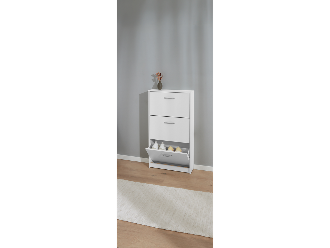 A white three-drawer shoe cabinet with a pull-out bottom drawer sits on a light wood floor against a gray wall.
