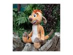 Timon plush toy sitting on a log amidst green and purple foliage.
