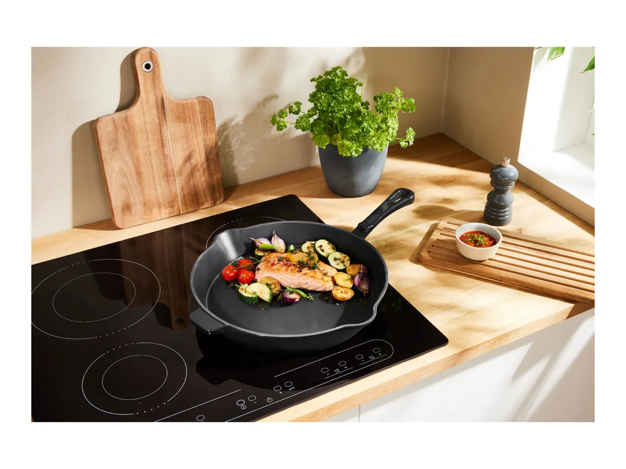 A black induction cooktop with a cast iron pan cooking salmon and vegetables.