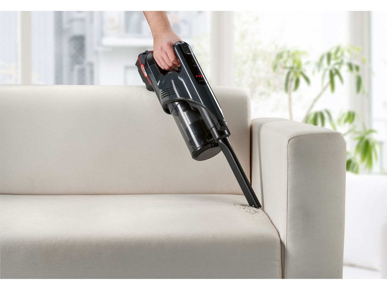 A hand holds a handheld vacuum cleaner cleaning a light beige couch.