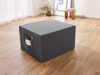 A dark gray ottoman with side pockets holding a book and remote control, on a wooden floor.