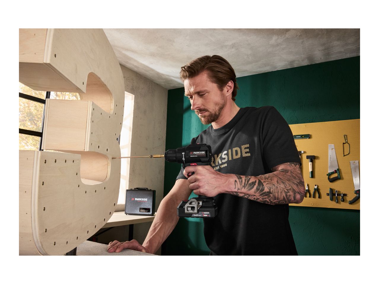 Man drilling into a wooden shelf with a cordless drill, surrounded by various tools.