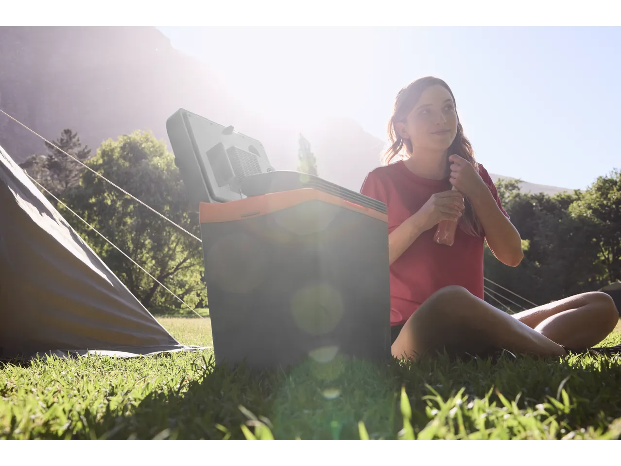 Woman sitting by a portable cooler and tent, holding a drink bottle in a sunny outdoor setting.