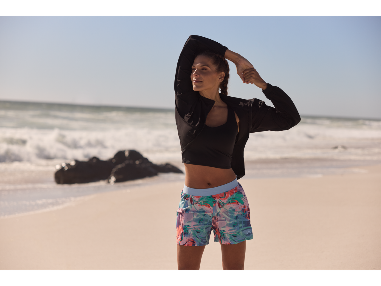 Femeie pe plajă cu sutien sport, jachetă și pantaloni scurți cu imprimeu.