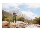 Man in fishing waders and t-shirt, holding a fishing rod in a river with mountains.