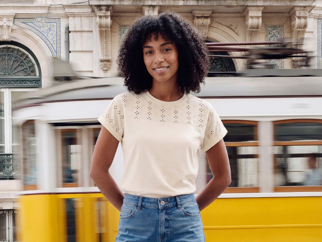 Woman in cream eyelet t-shirt and denim shorts, smiling in front of a moving tram.