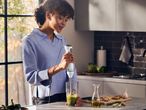 Woman blending green ingredients in a glass with an immersion blender in a kitchen.