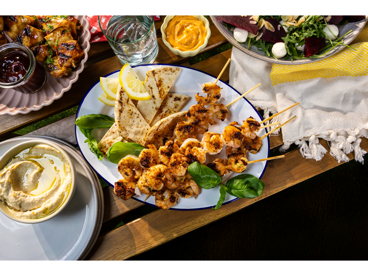 Grilled shrimp skewers, pita bread, hummus, BBQ chicken, and a beet salad on a wooden table.