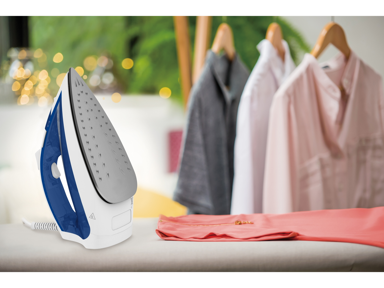 Fier de călcat cu aburi Tefal® pe o masă de călcat, cu haine în fundal.