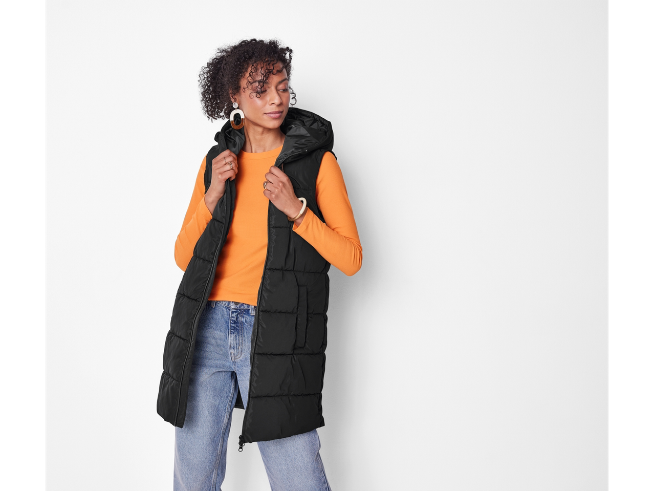 A woman in a black puffer vest, orange long-sleeve shirt, and light-wash jeans.