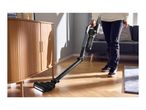 A person vacuuming a wooden floor under a cabinet with a cordless stick vacuum cleaner.