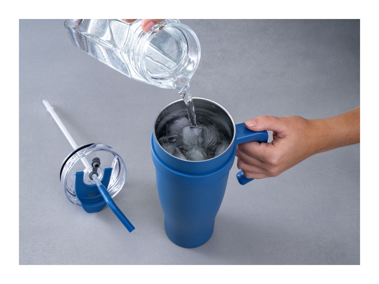 Water being poured into a blue tumbler with ice, next to its lid and straw.