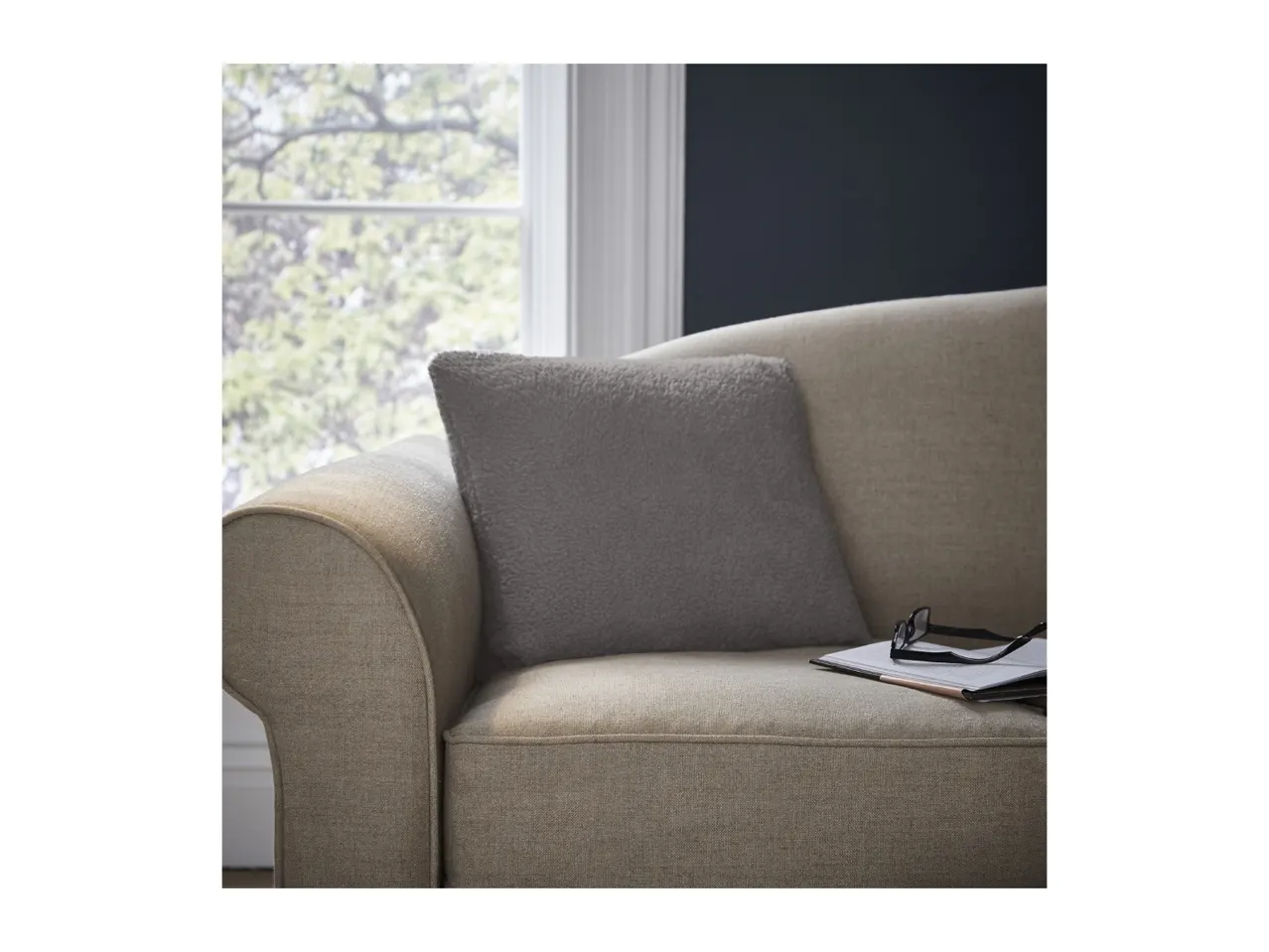 A cozy sofa with a grey cushion, glasses, and a book, by a window with green trees.
