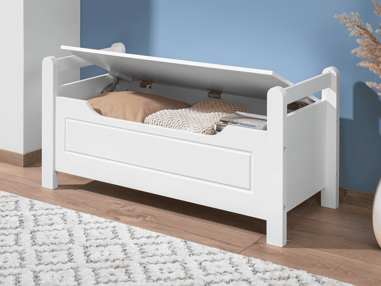 Weiße Truhenbank mit geöffnetem Deckel, Kissen und Decke, auf Holzboden mit Teppich.