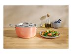 A peach-colored cooking pot with a lid, a plate of vegetables, olive oil, and a mortar and pestle on a wooden surface.