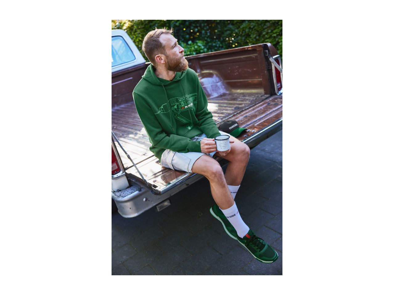 Man in a green Parkside hoodie and shorts, sitting on a pickup truck tailgate, holding a mug.