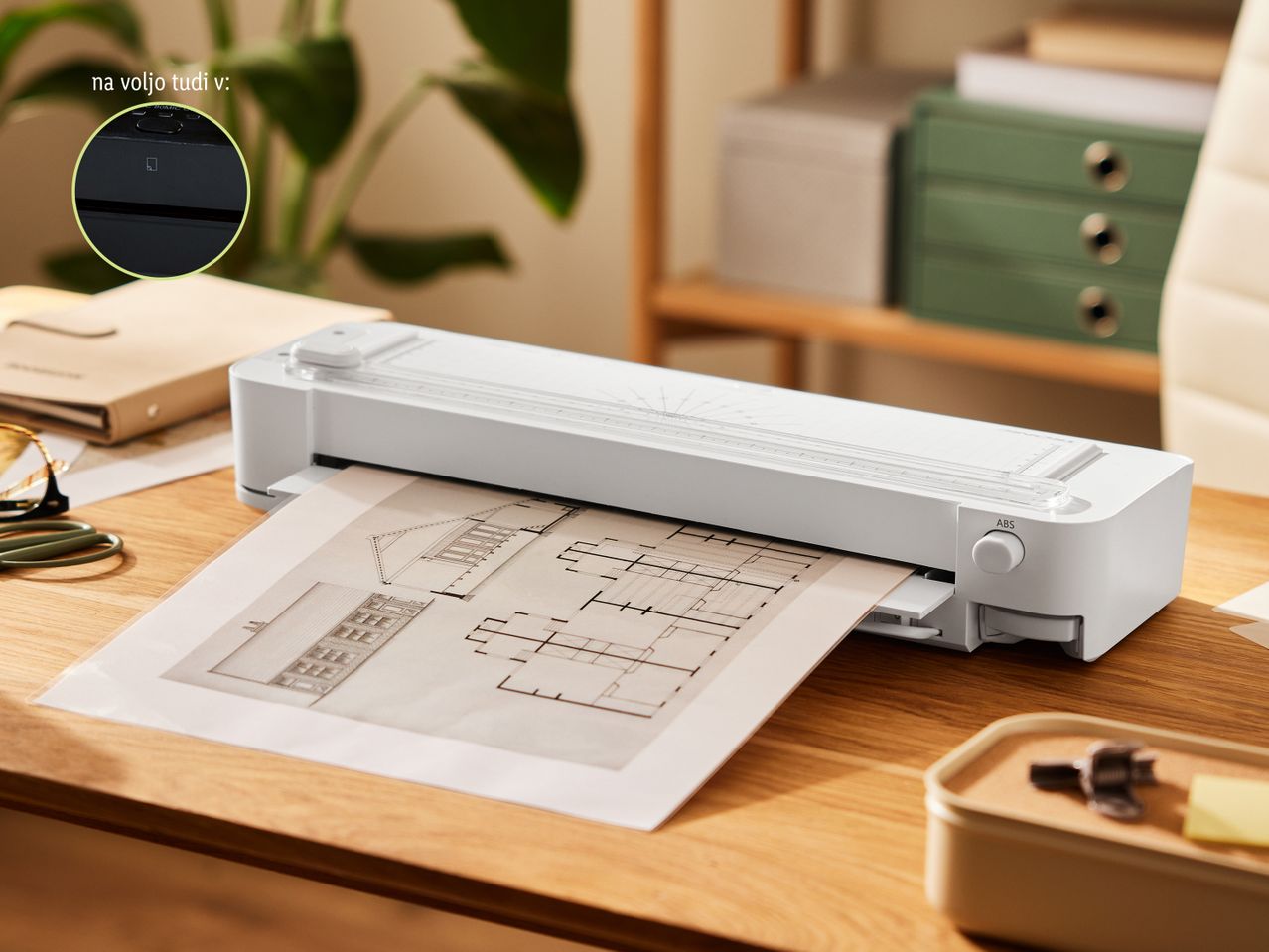 Beli laminator na leseni mizi, ki laminira načrt, z rastlino in pisarniškimi predmeti v ozadju.