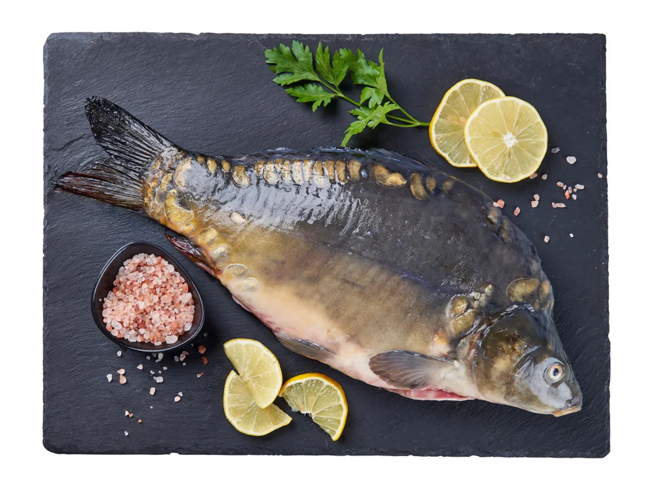 Svaiga vesela karpa zivs uz melnas šīfera dēļa ar citronu šķēlītēm, pētersīļiem un rozā sāli.