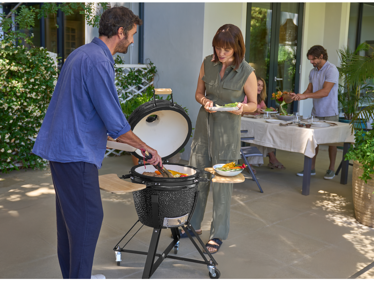 Man grilling vegetables on a ceramic barbecue, woman holding a plate of food.