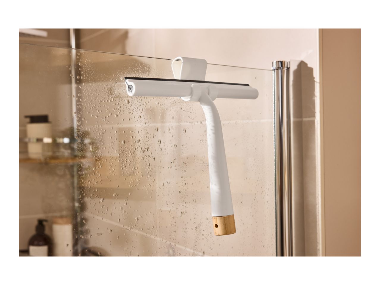 White shower squeegee with a wooden handle hanging on a wet glass shower door.