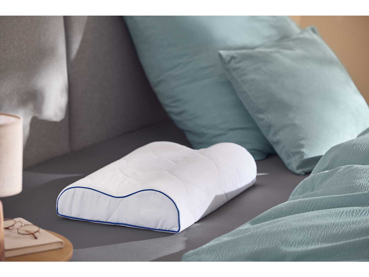 Ergonomic pillow on a bed with blue bedding and a lamp on a nightstand.