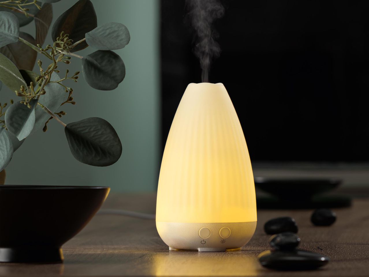 Világító aroma diffúzor gőzzel, fekete tál és eukaliptusz ágak mellett.