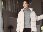 Frau in beiger Pufferjacke und grauem Rollkragenpullover, die aus einer Tür schaut.
