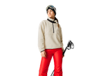 Woman in beige fleece, red ski pants, and ski goggles, holding ski poles.