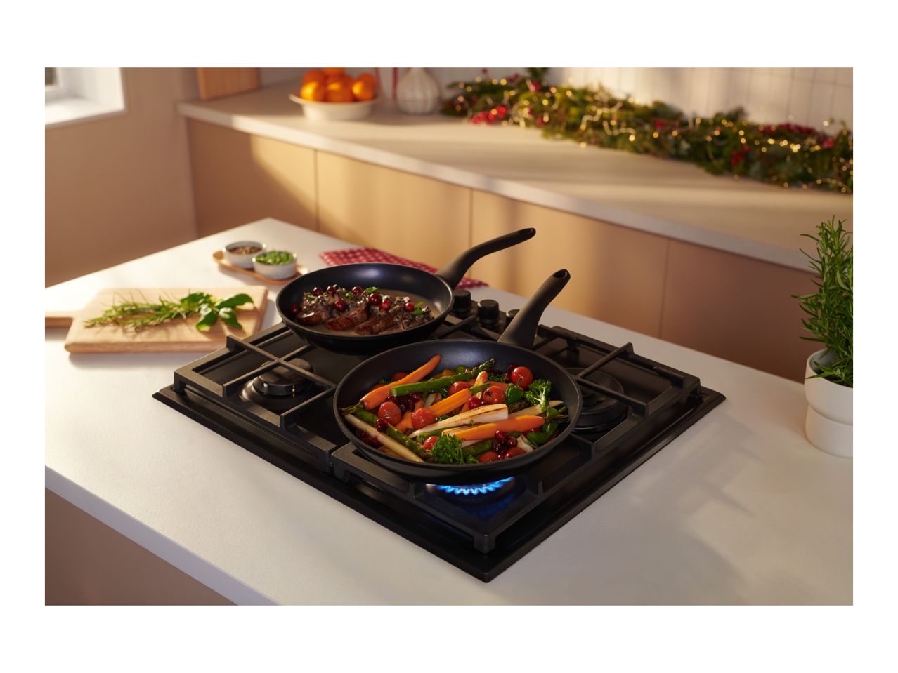 Two frying pans with food on a gas hob in a kitchen decorated for Christmas.
