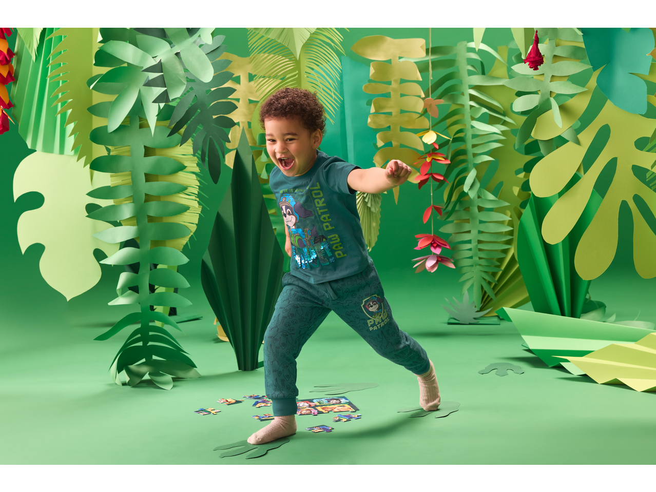 Criança de pijama verde com motivo de desenho animado, a brincar com um puzzle num cenário de selva de papel.
