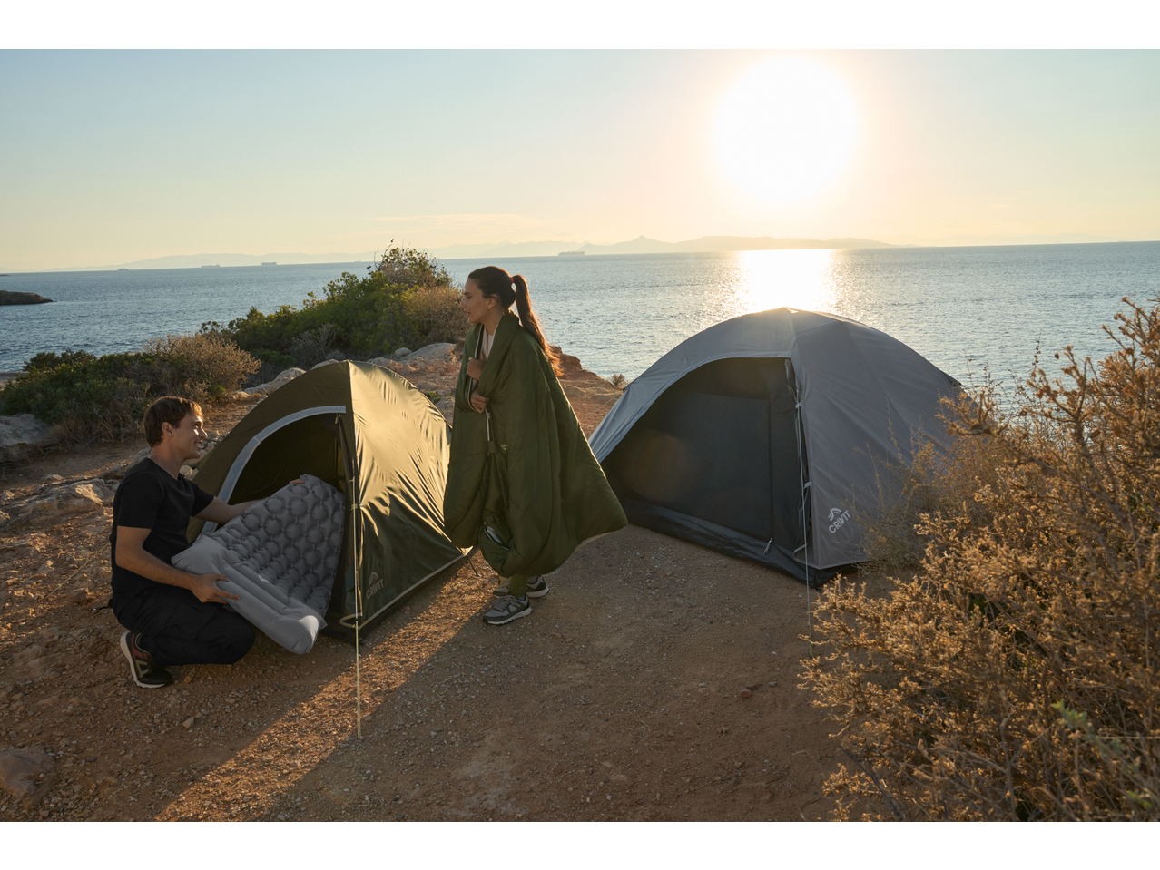 Two people camping by the sea with two tents and an inflatable sleeping mat at sunset.