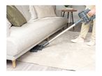 A person vacuuming under a light grey sofa with a cordless stick vacuum cleaner.