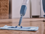 Моп за пръскане почиства дървен под, с видим резервоар за вода и синя подложка.