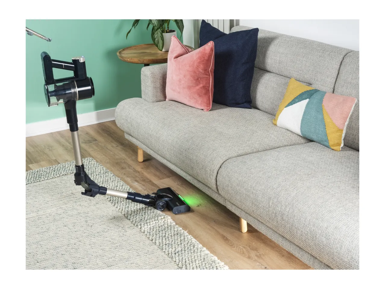 Cordless stick vacuum cleaner cleaning under a grey sofa with colorful cushions.