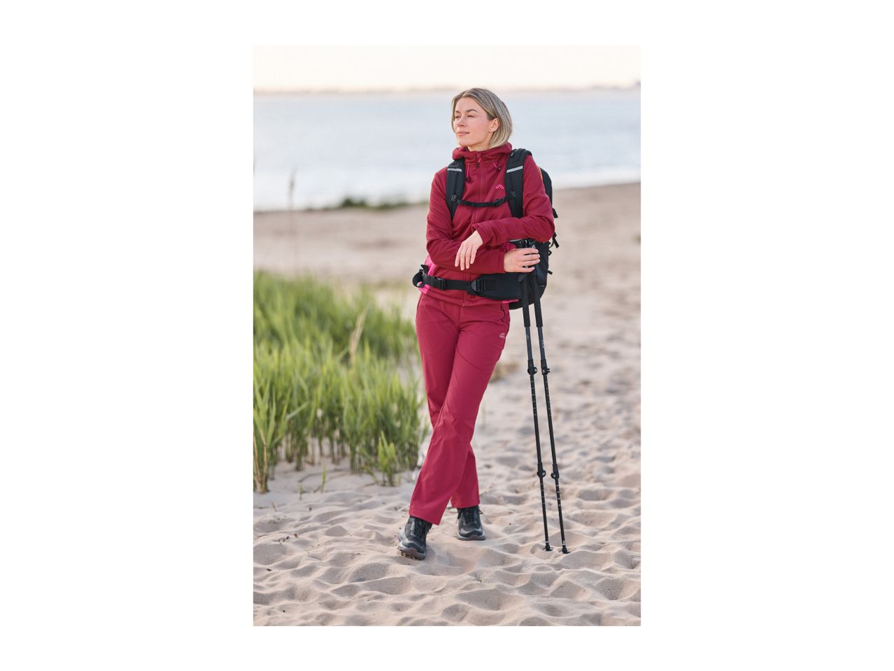 Woman in a red hiking outfit with a backpack and trekking poles on a sandy beach.